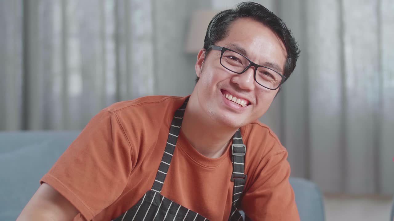 집에서 스프레이로 테이블을 청소하는 동안 카메라를 향해 미소 짓는 아시안 남성 집주인의 클로즈업