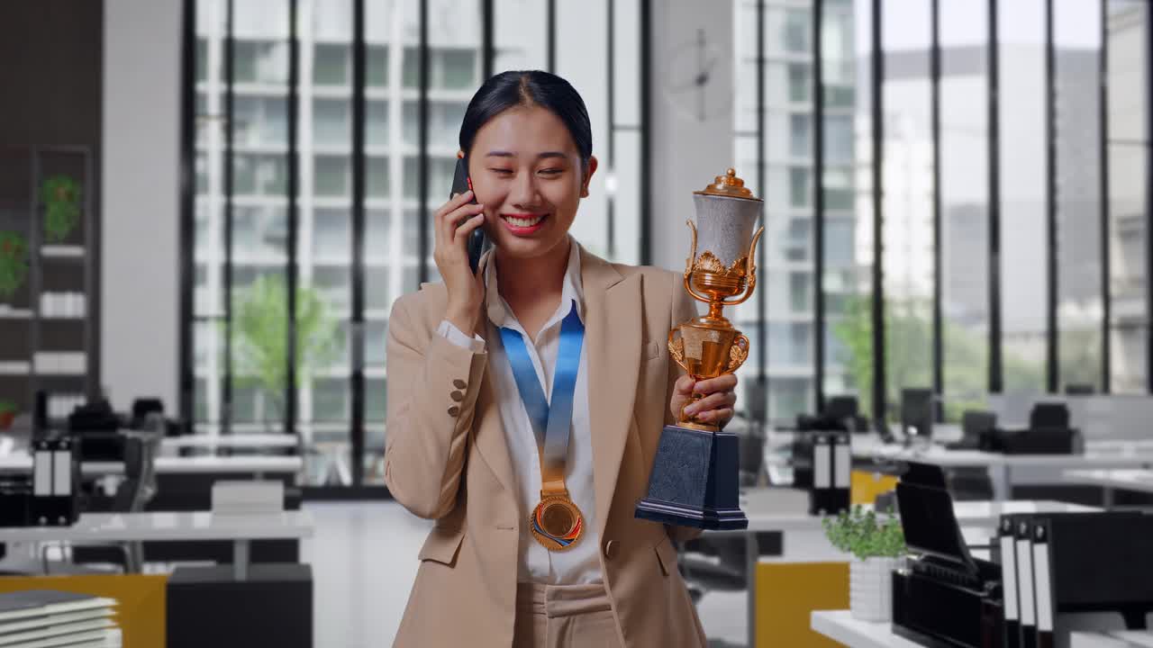 Asian Business Woman In A Suit With A Gold Medal Holding A Gold Trophy And Talking On Smartphone In The Office