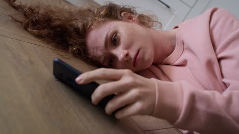 Sad-young-caucasian-woman-lying-on-floor-and-checking-mobile-phone