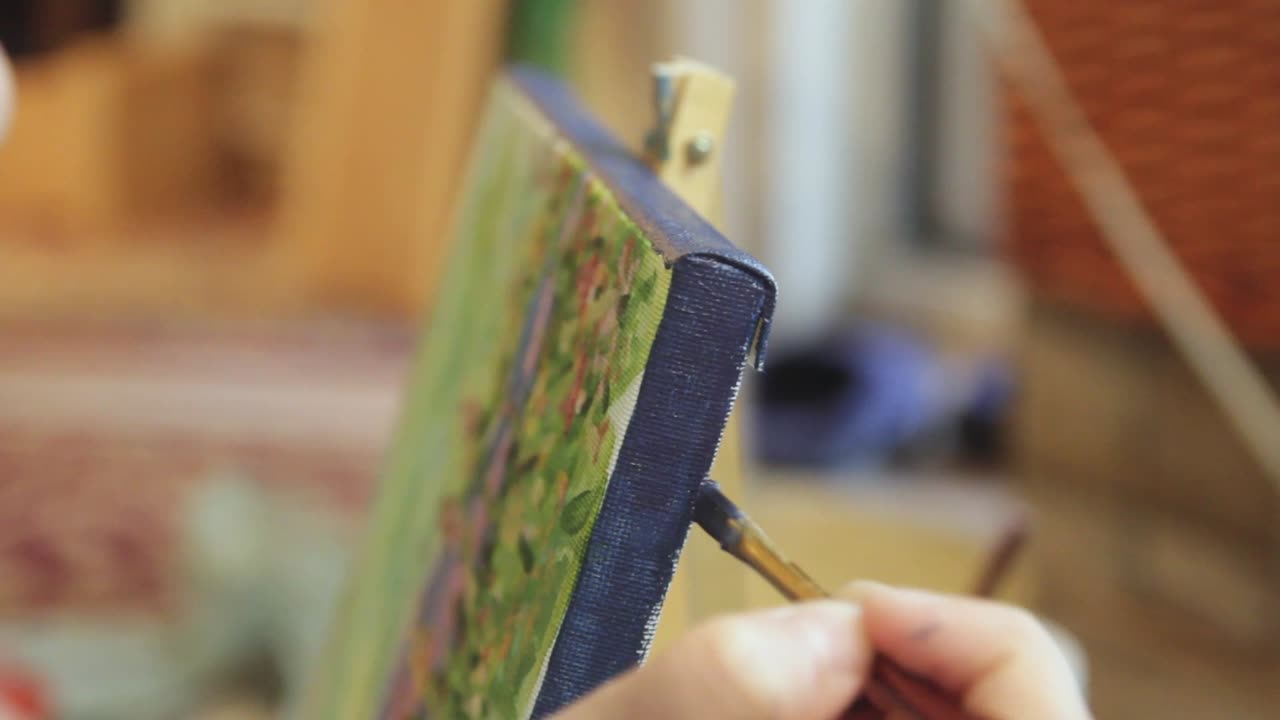 Female hand holding paintbrush painting the edge of canvas to finalise the artwork in a studio environment at daytime.