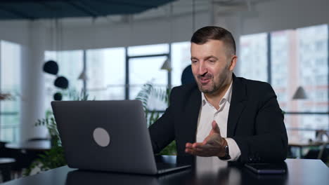 business meeting via laptop. businessman talking and smiling while talking before the laptop. spacious office in blur at the background.