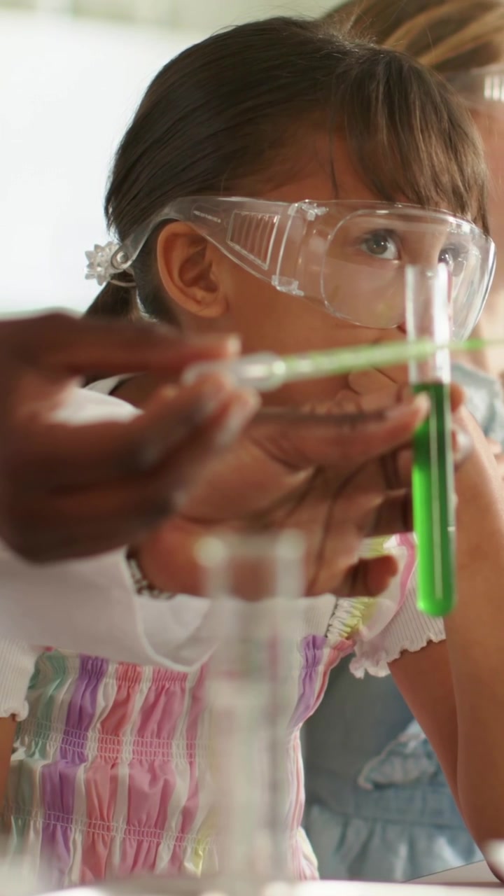 Vertical video: Bringing green tube pipette, adult showing girl pink dress leaning at lab goggles