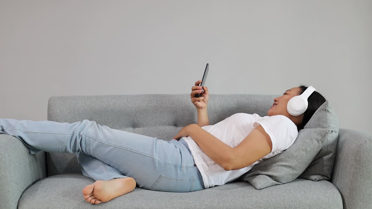 Asian adult woman using smartphone and scrolling social media while lying on couch at home and relaxing