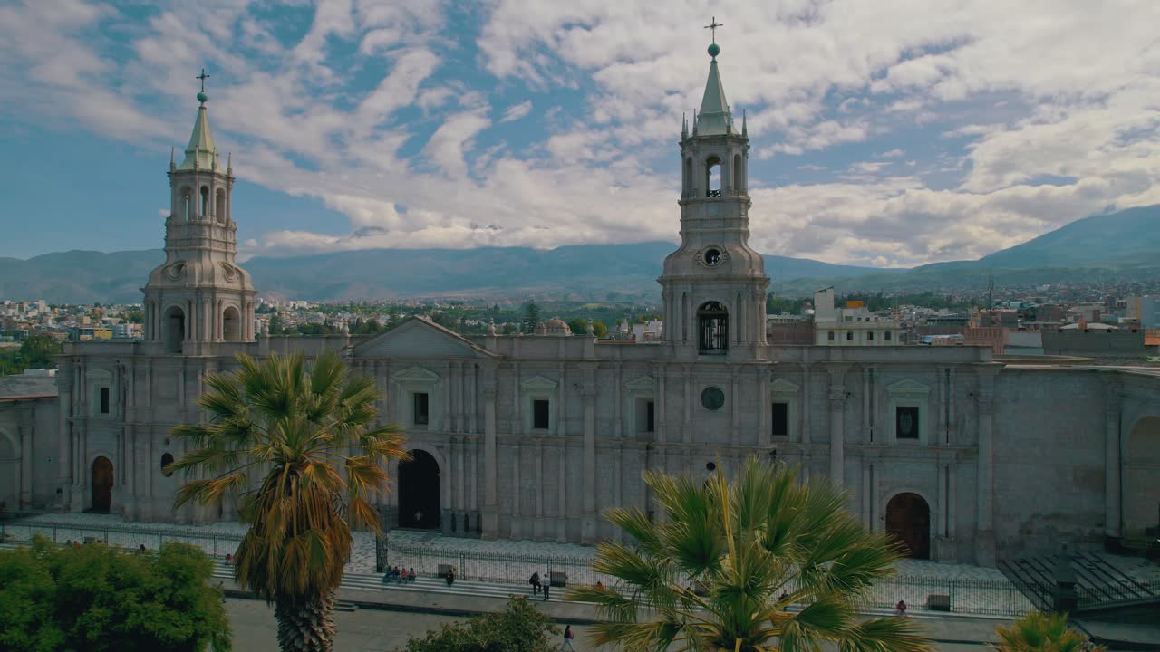 panorámica aérea de drones de izquierda a derecha que muestra la catedral de arequipa en un día nublado con luz solar intermitente, capturando a la gente en las calles y la vegetación local que decora la plaza