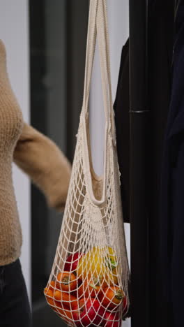 vertical video of woman getting home from food shopping hanging up coat and carrying bag of fresh vegetables indoors into kitchen 2