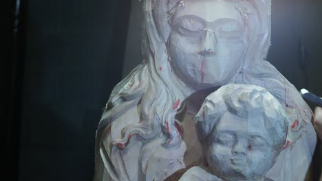 a detailed close-up of a wooden madonna and child sculpture in progress, showing tool marks and red chalk guidelines as the artisan refines the piece in a traditional woodworking studio
