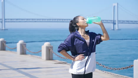 Ropa-Deportiva-Atractiva-Mujer-Deportiva-Bebiendo-Agua-Después-De-Hacer-Ejercicio-Trotando-Entrenamiento-Físico-En-Ropa-Deportiva-De-Pie-En-La-Orilla-Del-Mar-Cerca-De-La-Vista-Del-Puente