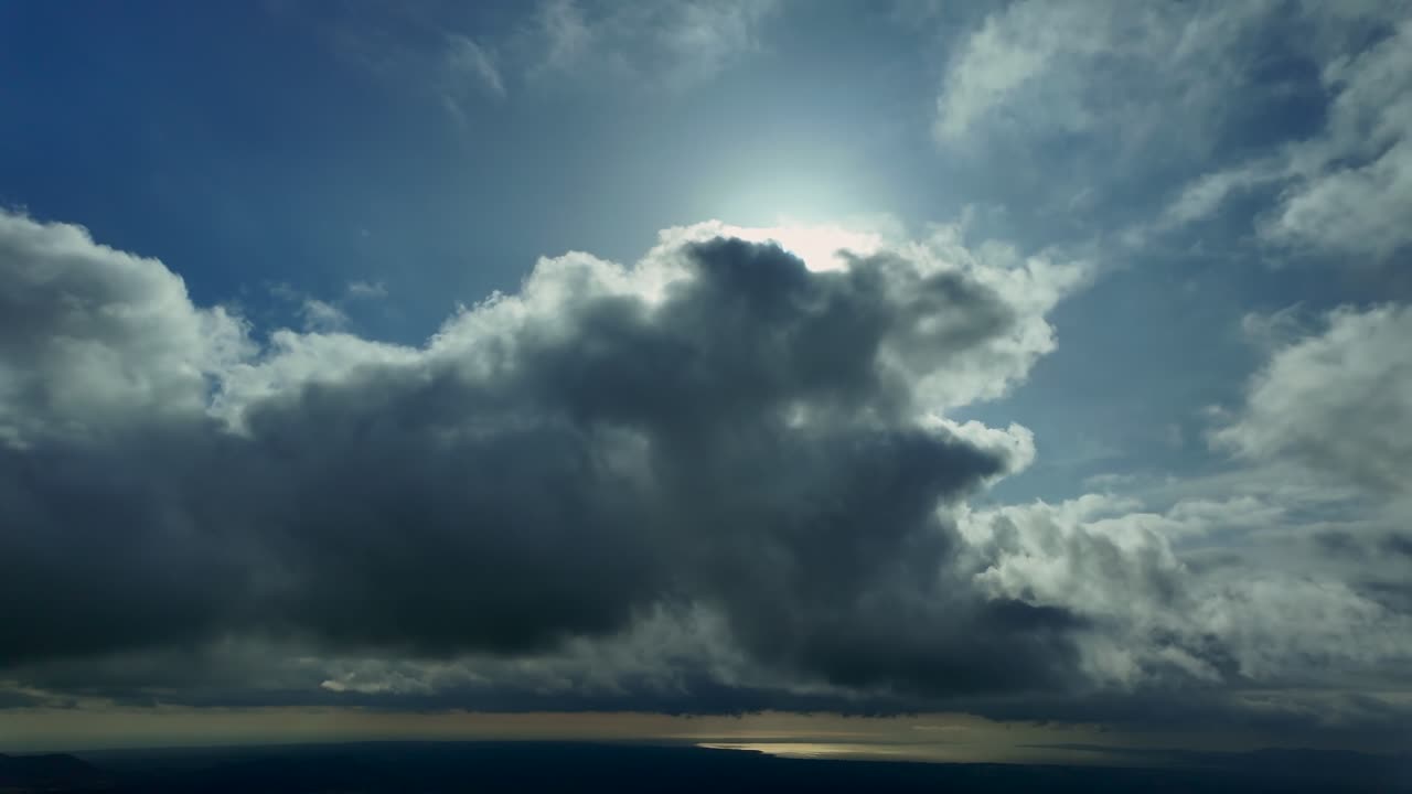 En luftbåren hyperlapse taget fra et jetflys cockpit, mens det flyver under et lag af brudte, vatagtige skyer med en strålende, tilsløret sol ovenover, og havet i baggrunden.