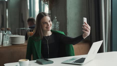 Una-Bella-Joven-Sentada-En-Un-Elegante-Restaurante-Vestida-Con-Traje-De-Negocios-Se-Toma-Una-Selfie-En-Su-Teléfono-Inteligente-Con-Una-Sonrisa