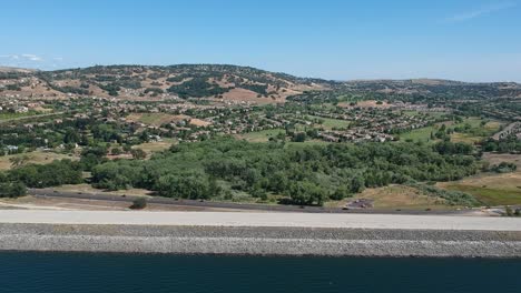 drone footage over a turquoise green lake and shoreline, showing a highway with cars, and a lush hillside with trees, fields and a town spread out