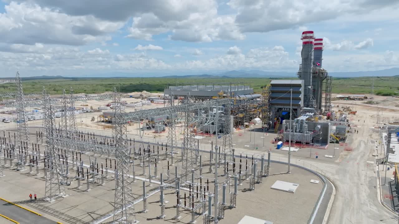Aerial view of large power plant with electrical substation in foreground, transmission towers, power lines, and smokestacks. Drone ascending