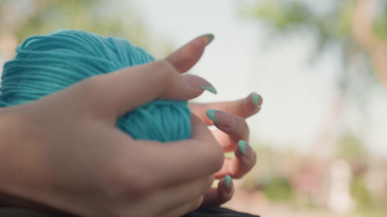 Close up of hands with turquoise nail polish holding ball of blue yarn. person manipulates soft material revealing texture details and gentle movement against outdoor backdrop with blurred