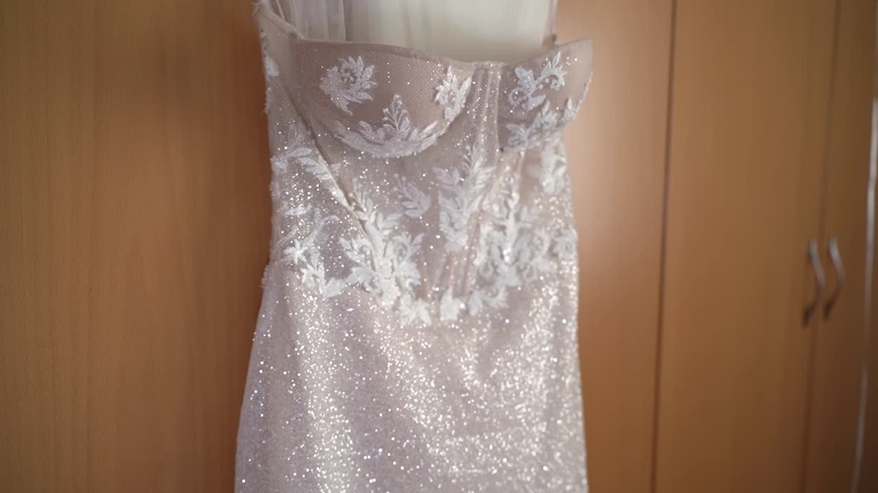 Un abito da sposa è esposto su una parete all'interno di una stanza. La luce risplende sull'abito, mettendone in risalto il design. L'ambientazione suggerisce i preparativi per un'occasione speciale.