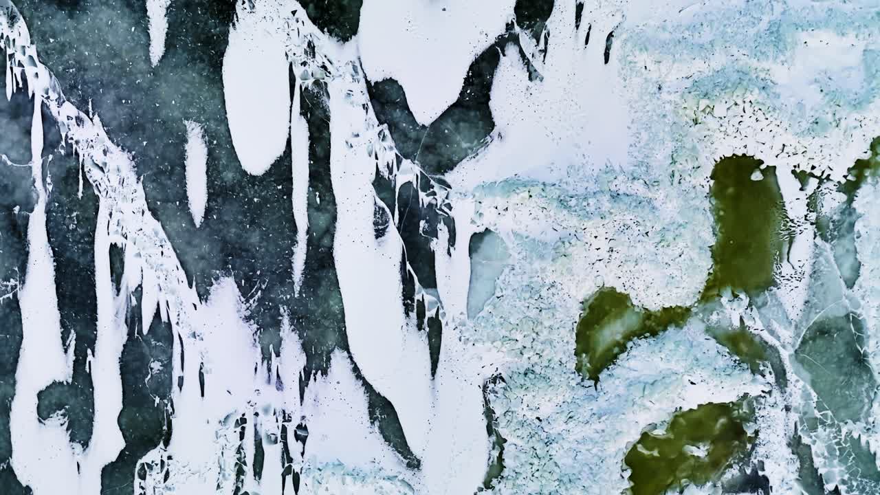 Vista dall'alto con drone del ghiaccio congelato del Mar Baltico con lunghe strisce bianche di neve, superficie screpolata blu-grigia e vivaci chiazze di alghe verdi che formano sorprendenti motivi invernali astratti