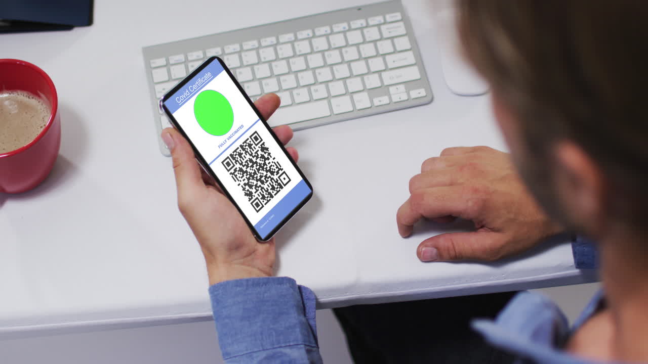 Man at desk holding smartphone with covid vaccination certificate and qr code on screen