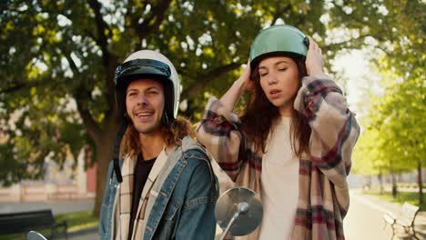 close-up shot of a brunette girl in a plaid shirt preening herself, looking into the side window of a moped, putting on a green helmet and looking at her boyfriend in a denim shirt who smokes a cigarette in a white helmet while sitting on a motorcycle in a sunny city on the street