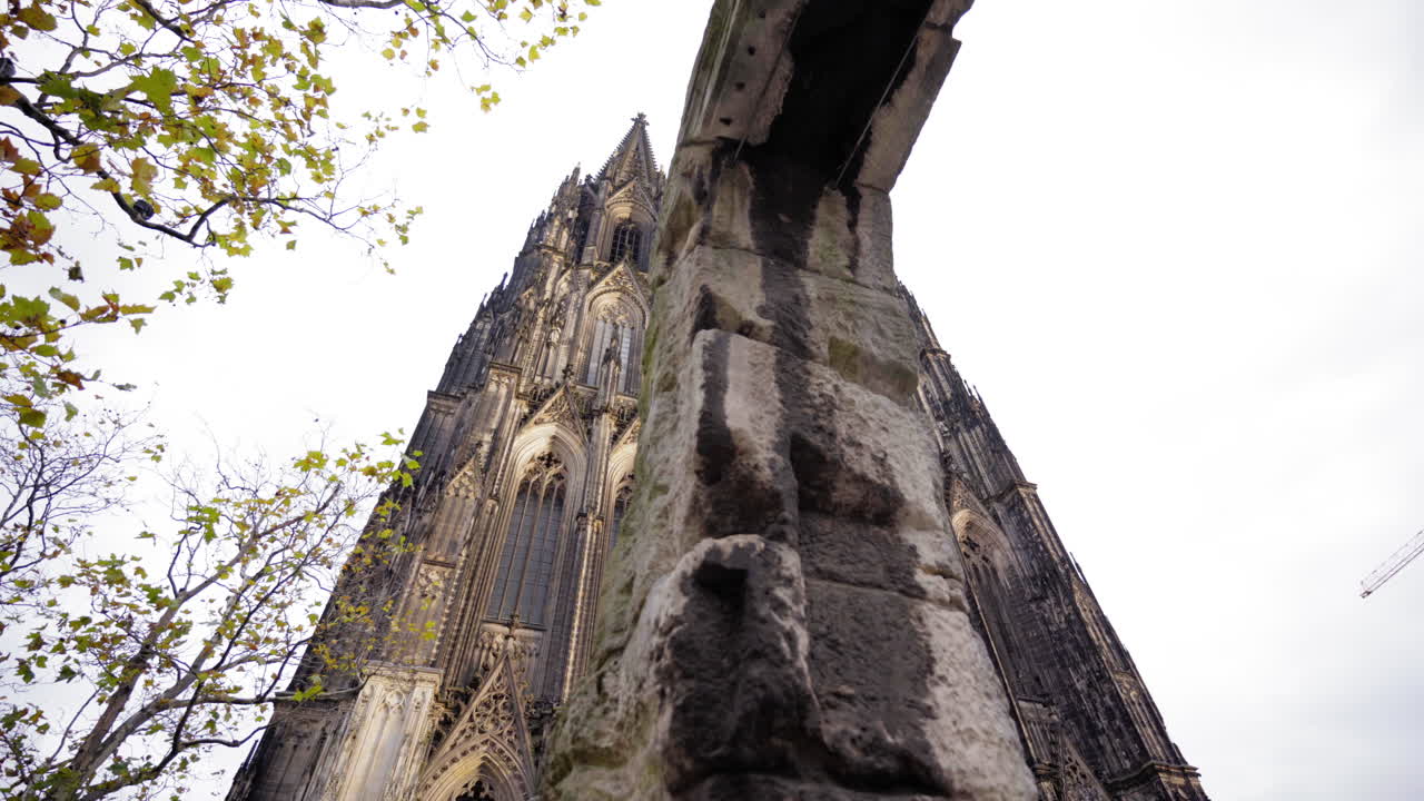A majestic view of the Cologne Cathedral's twin spires framed by autumn leaves and ancient stonework, showcasing its intricate Gothic architecture under a cloudy sky