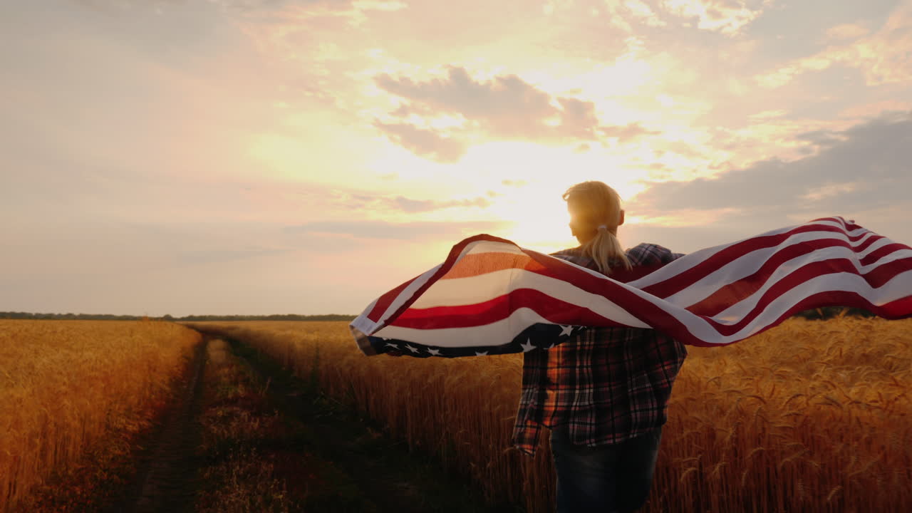 Una donna con una bandiera USA corre attraverso un campo di grano sotto i raggi del sole al tramonto.