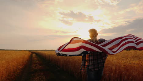 una mujer con una bandera de estados unidos atraviesa un campo de trigo bajo los rayos del sol al atardecer