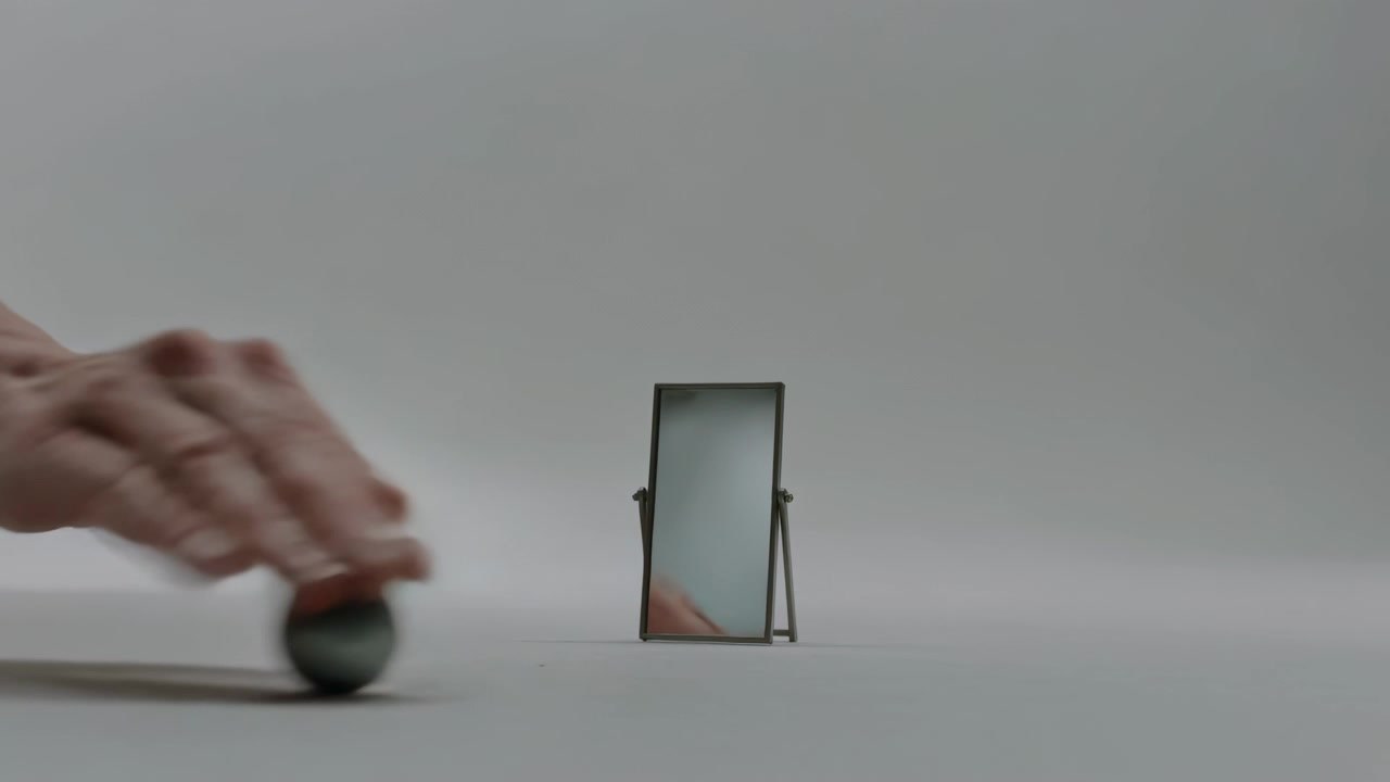 Una mano umana nuda entra da sinistra, spingendo una sfera d'argento che rotola su un piano grigio, oltre uno specchio.
