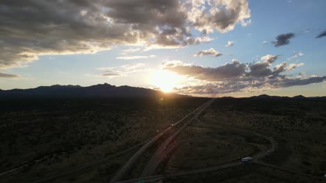 aerial view drone panning around highway exit ramp with sun setting behind mountains in the background, light blue sky and large clouds, lens flare