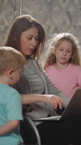 mother shows curious kids pictures on laptop sitting in wheelchair. young woman with disability enjoys educating little son and daughter at home