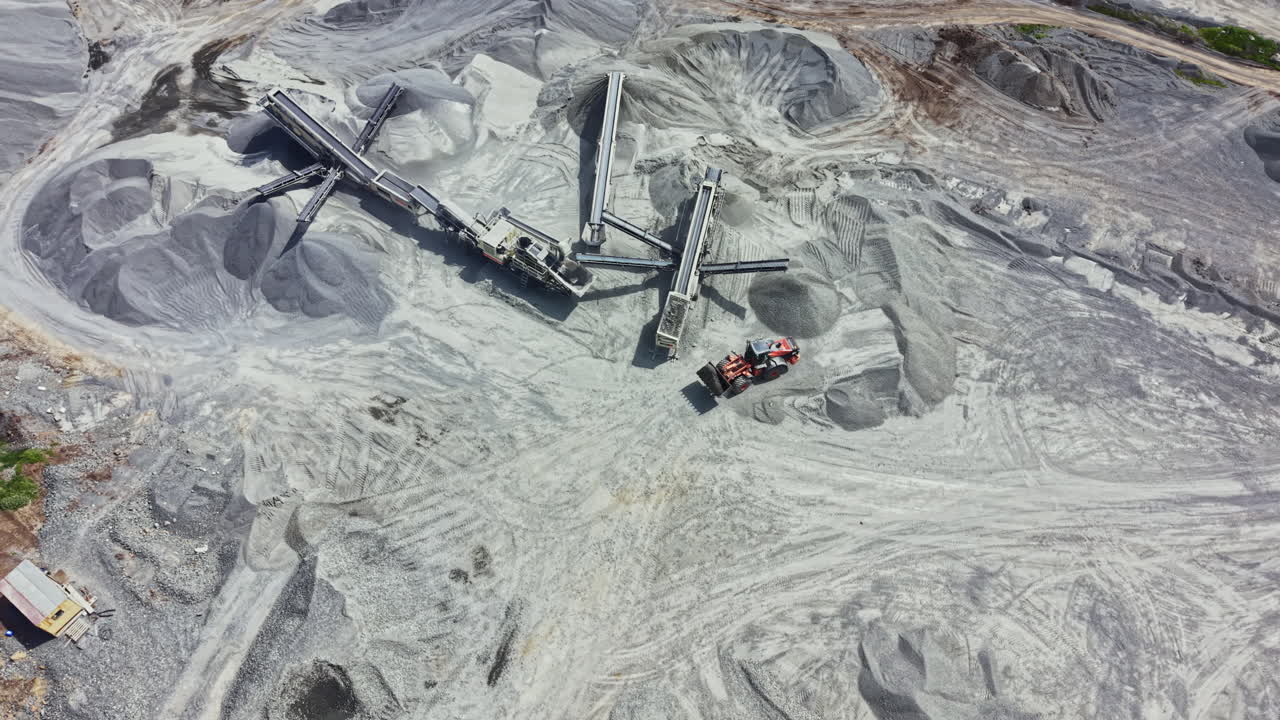 Operations and machinery at a granite quarry during daytime showing trucks, conveyor belts, and granite piles in a work site