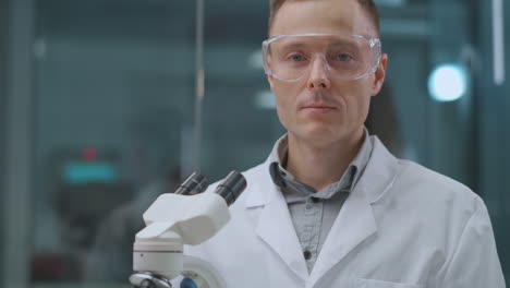 retrato de un técnico de laboratorio confiado, un hombre en la sala de trabajo con gafas de protección de pie cerca de un microscopio, un químico o un profesional de la salud