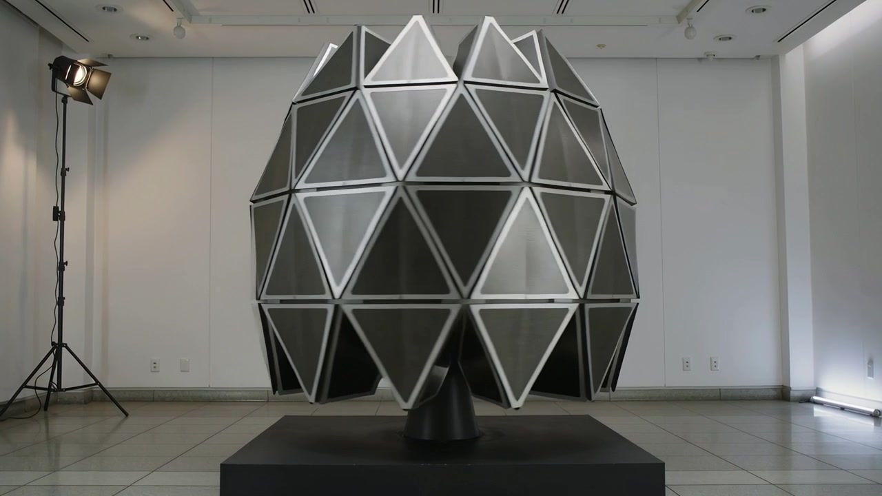 Attivazione del motore, scultura a sfera geodetica che incerniera i pannelli su un piedistallo in galleria, rivelando una corona