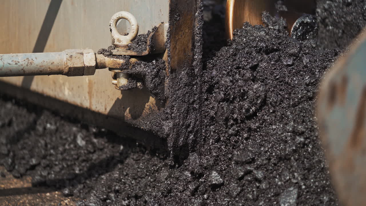 Close-up of hot asphalt pouring out the paver machine to the road. Asphalt paver machine during road construction. Heavy truck laying out black bitumen.