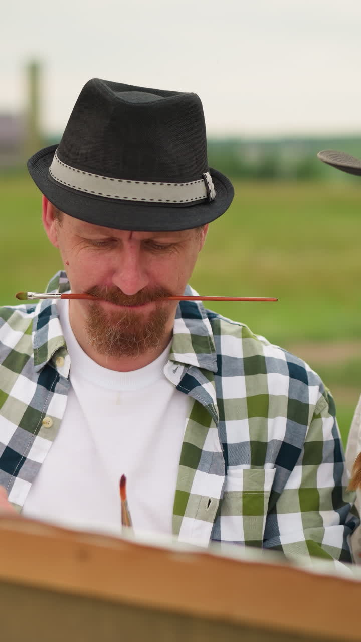 A scene of an artist focused on his work, holding a paintbrush in his mouth while wearing a checkered shirt. Beside him, a woman in a white dress and hat stands, smiling as she gently touches his nose