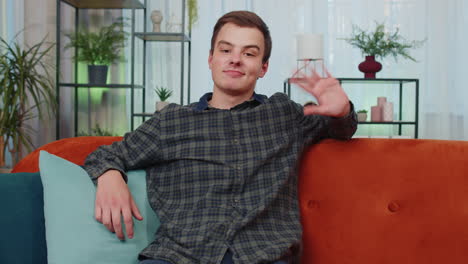 Teenager-man-smiling-friendly-at-camera,-waving-hands-gesturing-hello,-hi,-greeting-at-home-on-couch