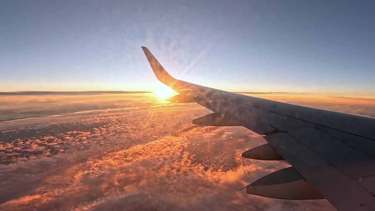 Video from an airplane window seat showing the aircraft wing against a stunning sunset above a thick cloud layer. Golden and orange tones illuminate pink clouds a beautiful scenery