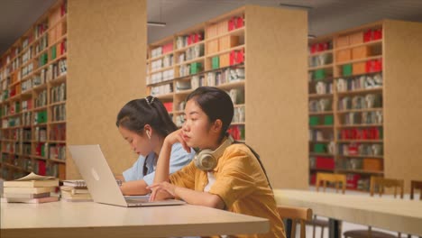 asian woman student with headphones are bored typing on a laptop while sitting with her classmate studying on a table in the library