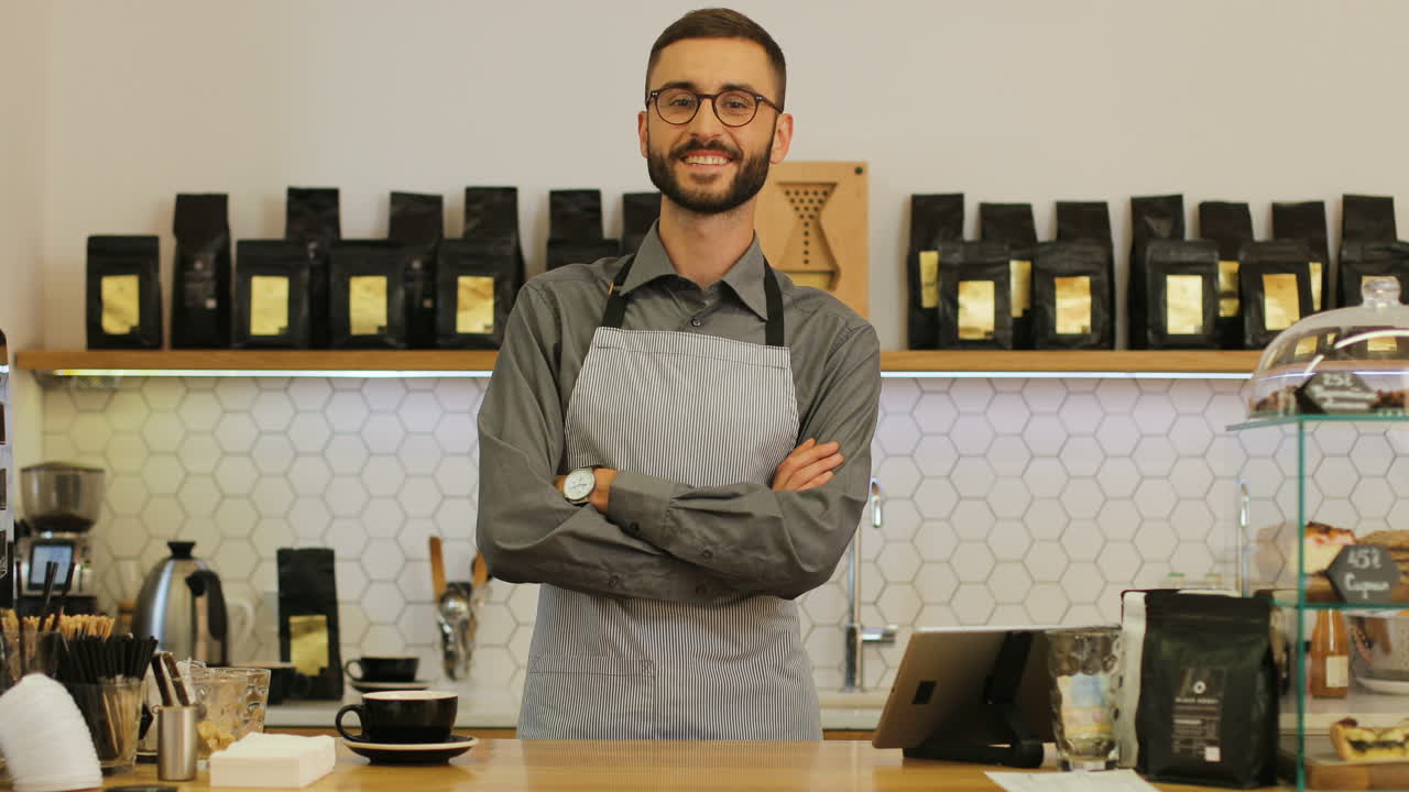 vista de primer plano de un barista masculino feliz usando gafas en la cafetería posando y sonriendo a la cámara con los brazos cruzados