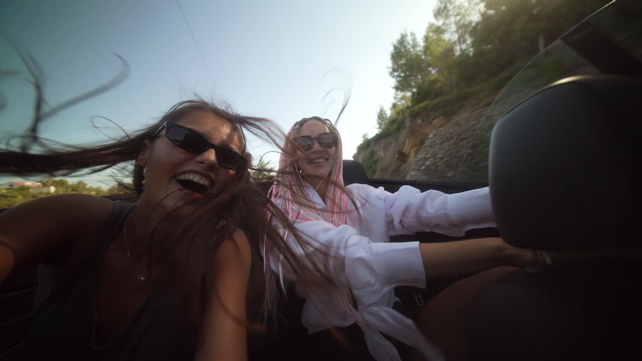 Two joyful women on a convertible road trip, hair blowing in the wind, taking selfies