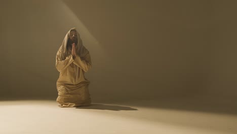 Studio-Portrait-Of-Kneeling-Man-Wearing-Robes-With-Long-Hair-And-Beard-Representing-Figure-Of-Jesus-Christ-Praying-