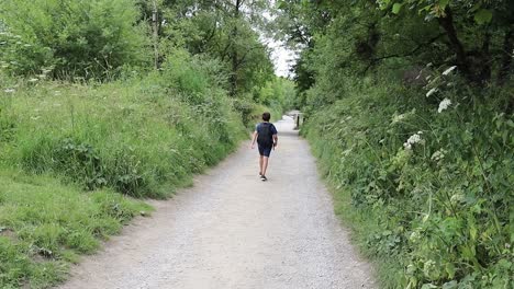 Young-boy-walking-on-a-footpath-through-a-treelined-area,-alone-on-a-bright-summer-day