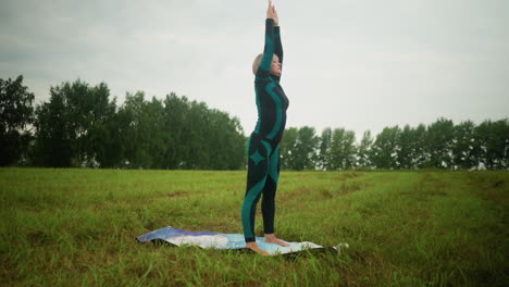 side view of woman with hands raised up on yoga mat, slowly bringing them down, bending close to her knees, and folding her arms, twisting left and right, in vast grassy field under a cloudy sky