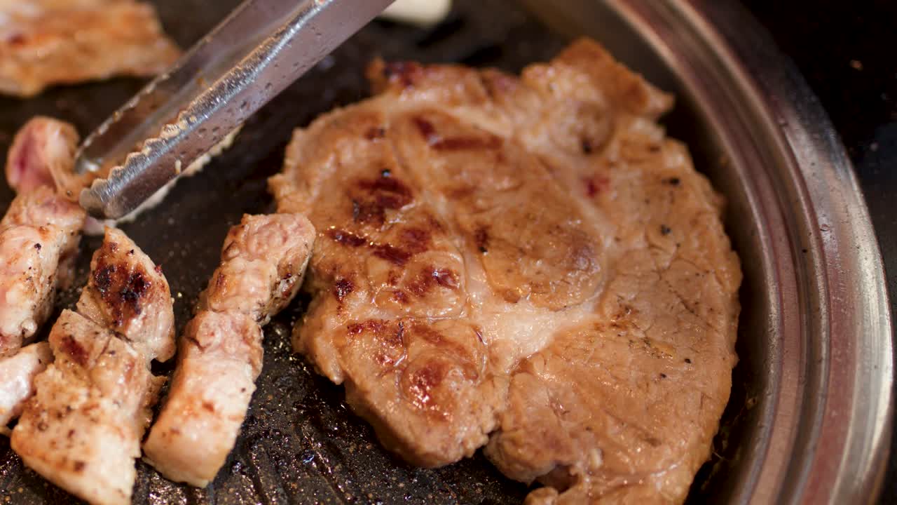 Tongs turn pork belly and steak on a hot Korean barbecue grill under bright lights