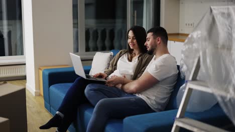 Eine-Hübsche-Schwangere-Frau-Und-Ein-Mann-Benutzen-Einen-Laptop-Und-Sitzen-Auf-Dem-Sofa-In-Einem-Neuen-Wohnzimmer