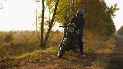 vista posterior de un hombre con casco y chaqueta de cuero acercándose a su bicicleta y arrancando el motor mientras estaba de pie en la carretera en un día soleado de otoño