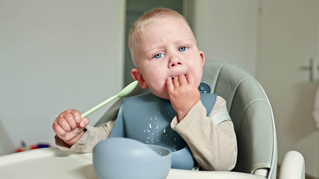 Cheerful toddler boy eats milk porridge with a spoon while sitting in a high chair. Child eats with a spoon from a silicone plate. Safe silicone dishes for children