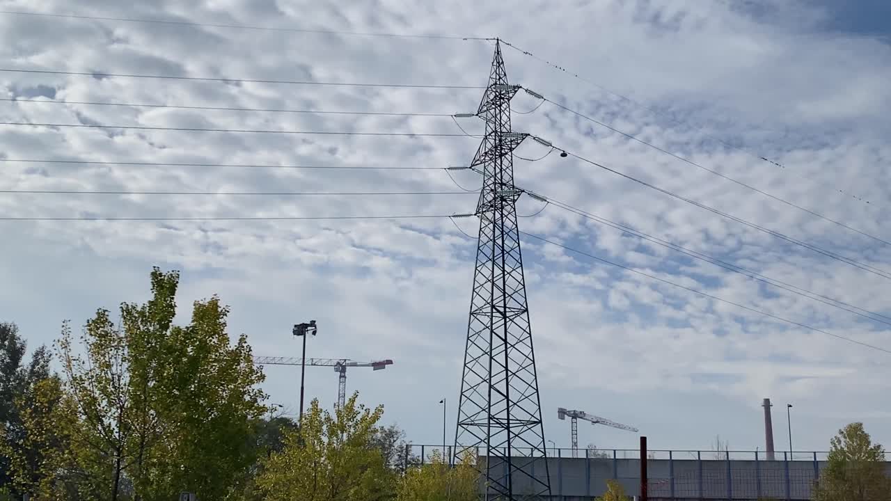 An electricity pylon against a cloudy sky with power lines and distant industrial elements