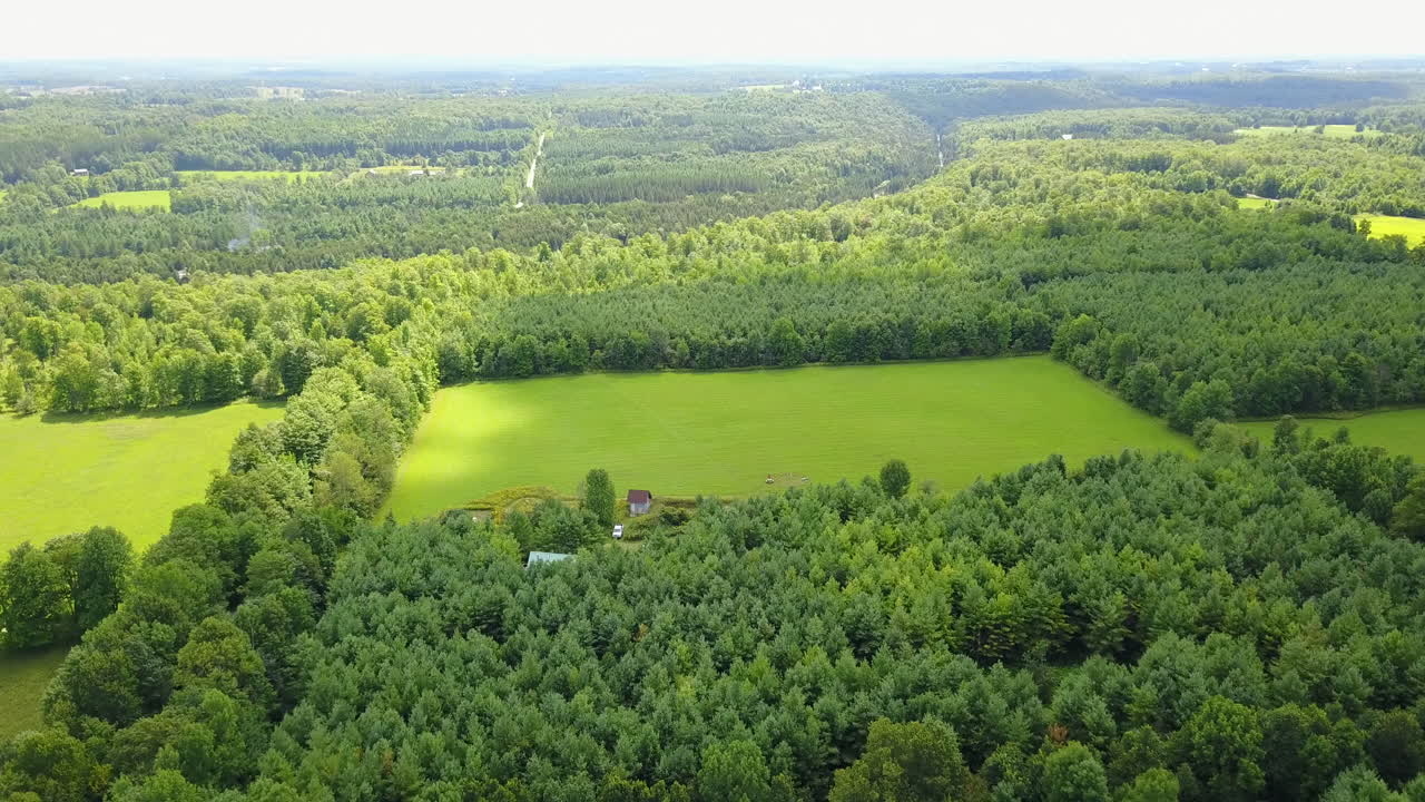 Dron letící nad rozlehlým lesem směrem k malé zemědělské usedlosti.