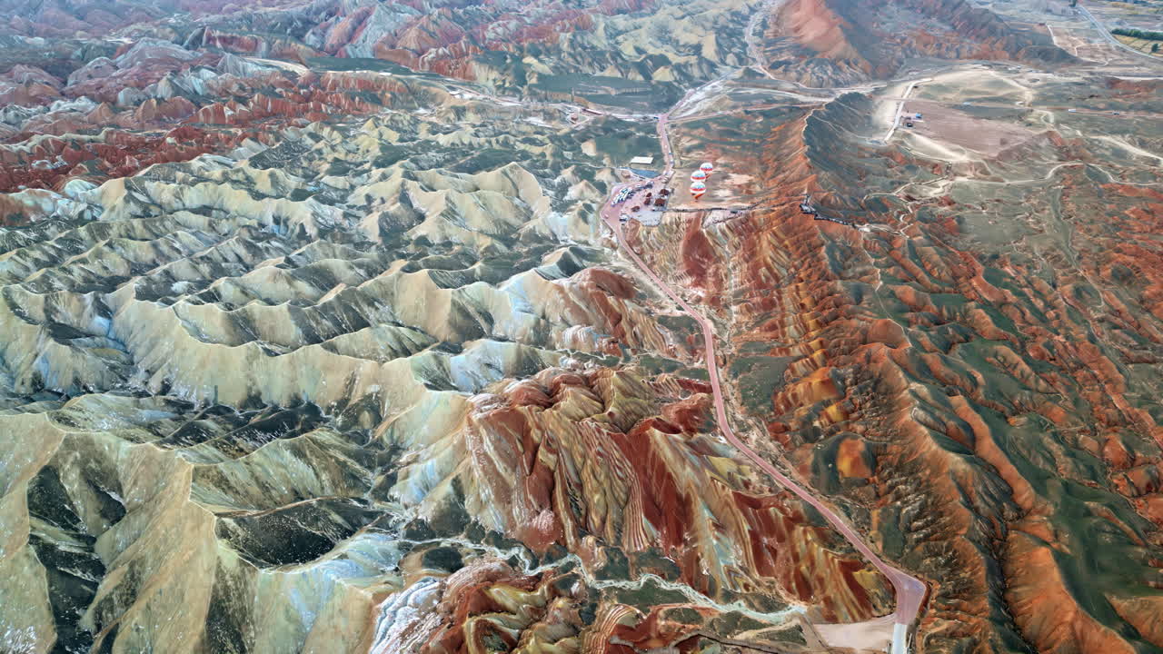 Dronekuva Zhangye Danxia National Geoparkista, Zhangyesta, Gansusta, Kiinasta. Mutkitteleva aavikkotie leikkaa moniväristen raidallisten kukkuloiden läpi, samalla kun kuumailmapallot valmistautuvat lähtöön näköalapaikan lähellä