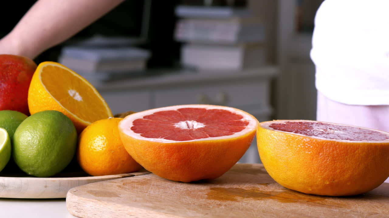 Woman slicing grapefruit in two parts on a wooden board. Fruits on the background. Slow motion