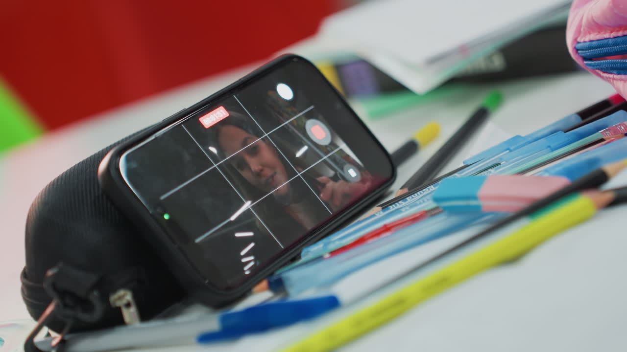 Smartphone apoyado en un estuche mostrando a un estudiante en una videollamada, bolígrafos, lápices y un cuaderno de colores esparcidos por el escritorio, configuración de estudio informal, ambiente de aprendizaje a distancia con un ensayo concentrado y sincero
