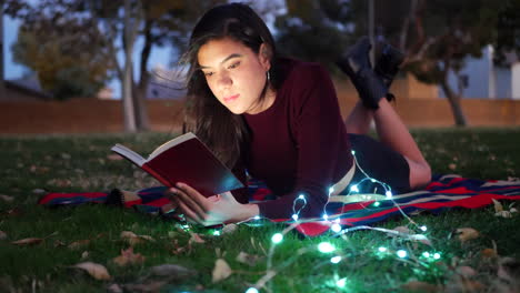 una joven estudiante hispana leyendo un libro al aire libre al atardecer con luces brillando en la oscuridad nocturna