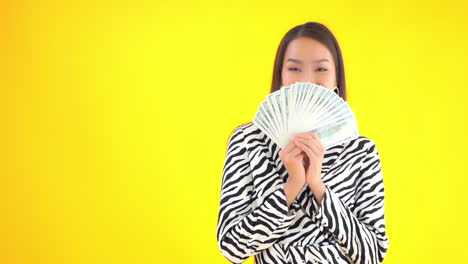 pretty playful asian girl holds up cash money, yellow background slomo